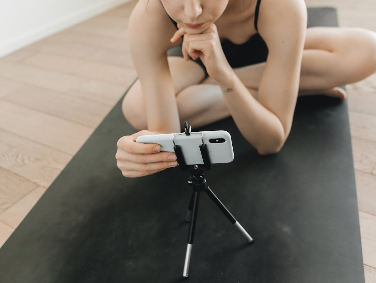 services-03 A woman in activewear vlogs using a smartphone and tripod while seated on a yoga mat indoors.